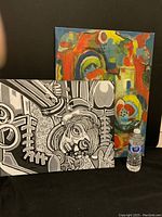 Two paintings displayed against a black background with a water bottle for scale. The front painting is monochrome abstract figurative in black, grey and white. The painting behind is colorful abstract with visible damage.