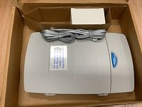 Top view of a gray Nortel phone system unit with attached cable, placed inside an original open box.