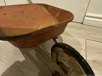 Front angle view showing the orange metal tray with wear and rust, part of wooden handles and rubber-tired metal wheel in cream paint with oxidation.
