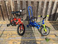 Two boys bikes shown side by side, one red and one blue, both with removable training wheels, positioned on a wooden deck.