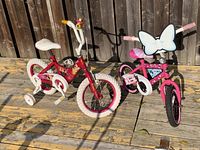 Two children's bicycles side by side on a wooden deck, showing the red bicycle with cream details and the smaller pink bicycle with a white bow on handlebar front.