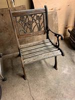 Original smaller size wooden bench with cast iron sides and decorative metal backrest showing leaf and vine pattern, situated on concrete floor.