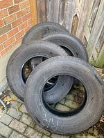 Four used Bridgestone Dueler tires stacked outdoors on a brick paved surface near a wooden fence and brick wall, showing overall condition and quantity.