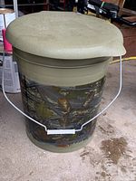Side view of the pail toilet showing the camo pattern on the bucket, green lid, and white plastic handle grip.