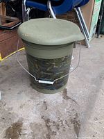 Green and camouflage plastic pail with a green toilet seat lid on a concrete floor with chair legs in background.