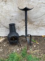 Full view of tall cast iron candle holder with spiral design and mini chiminea side by side on dirt ground against wood background.