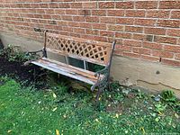 Front and side view of cast iron outdoor bench showing weathered wood and cast iron frame with lattice backrest design, placed outside against a brick wall on grass.