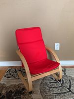 Child's lounger chair viewed at an angle showing the curved wooden frame and red fabric upholstery.