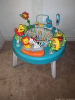 Full view of the Fisher Price infant exersaucer showing the circular blue base, fabric seat with multi-colored geometric pattern, and several attached toys including giraffe, lion, zebra, flower with bee, and other animal and activity toys.