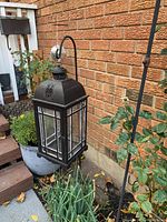 Black metal framed lantern with glass, hung on curved metal hanger outdoors, near plants and brick wall.