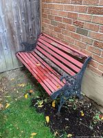 Full view of garden bench showing long wooden slats and cast iron ends with floral design. Bench placed outside on soil with brick wall background.