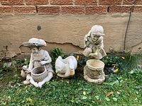 Wide view of three ceramic lawn ornaments positioned on grass against a wall, showing two child figures and a goose figure.