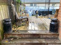 Two black milk cans placed outside on a small wooden platform against a fence.