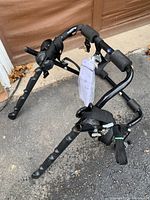 Bicycle carrier rack lying on the ground showing frame, padded foam arms, straps, and attached tag with model name.