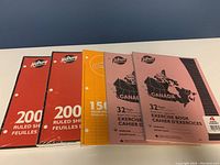 Photo showing 4 packages: two red packages with 200 ruled sheets each, one orange package with 150 ruled sheets and two pink exercise books.