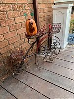 Front angled view of vintage iron bicycle plant holder showing multiple pot holders and front basket, placed on porch deck.