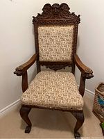 Full view of the antique carved wood cushioned chair showing upholstered seat and back with patterned fabric, decorative nail head trim, and carved armrests and legs.