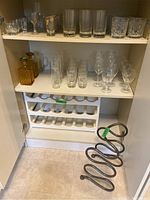 Photo of various glassware on cabinet shelves including shot glasses, wine glasses, and decanters. Also shows plastic and metal bottle racks below.