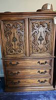 Front view of the dresser showing two ornate carved doors above three drawers with metal handles and a flat top with hats on it.
