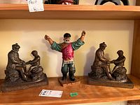 Photo showing the three figurines on a wooden shelf including two metal sitting figures and one painted standing figure with arms raised.