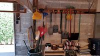 Garden tools on wall and inside bin including rakes, shovels, brooms, saws, and dustpan.