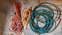 Photo showing green garden hose coiled state with orange and yellow extension cords beside it on garage floor with leaves.