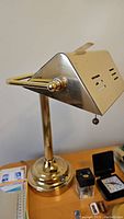 Brass-finished banker's type desk lamp showing the side profile of the rectangular shade with ventilation slots and pull chain switch.