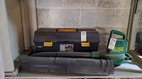 Side view of green and black Barracuda leaf blower next to a black toolbox (toolbox not included in the lot). Leaf blower placed on a shelf in garage.