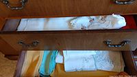 Wood drawer with folded linen napkins, placemats, and tablecloths in white, cream, blue, and yellow colors with embroidery and lace details.