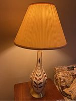 Single mid-century modern ceramic table lamp lit, showing the leaf motif on the body and pleated shade