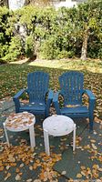 Two blue plastic outdoor chairs and two small white round plastic tables on outdoor ground covered with fallen leaves.