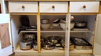 Open cabinet showing shelves with stacked pots, pans, mixing bowls, strainers, and baking trays