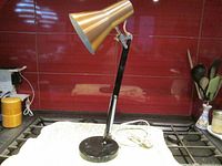 Full view of the black base and arm with copper conical lamp head positioned on a kitchen counter with a red tile backsplash.
