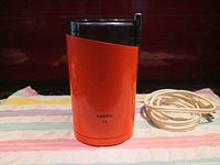 Front view of orange Krups coffee grinder model 75 with power cord coiled beside it.
