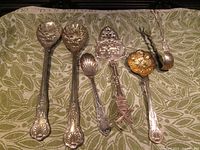 Wide view of six silver plated serving utensils arranged on patterned cloth showcasing intricate ornate designs and variety of shapes including scalloped spoons, pierced servers, salad servers, and a ladle.