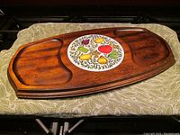Front angled view of brown wood cheese board with inset round tile with fruit designs and two recessed sections