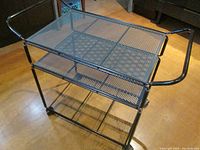 Bar cart shown from an angle illustrating its mesh shelves and black metal frame, highlighting the top shelf and side handle with caster wheels underneath.