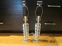 Pair of tall clear acrylic table lamps with metal fixtures, showing full height including finials, placed on a wooden surface against a dark background.