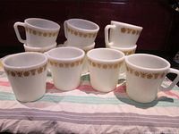 Front view of all 10 Pyrex coffee mugs arranged in two rows, showing the Butterfly Gold yellow and brown floral decal around the rim and diagonal handles.