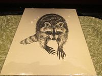 Full view of black and white wood cut print featuring a raccoon, sealed in clear plastic wrap on a green patterned surface.