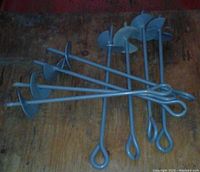 Photo showing eight metal screw-in ground anchors with a helix plate and an eyelet at the top, laid on a wooden surface.