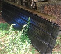 Four black metal sheets with ribbed profile, stacked outdoors on edge against a wooden structure and surrounded by vegetation.