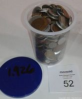 Clear plastic container filled with assorted world coins of various sizes and metals. Blue lid marked '1.1926' indicating weight.