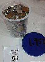 Clear plastic container filled with assorted world coins of various sizes and colors, blue lid beside it marked '1.950'.