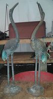Two metal stork sculptures standing side by side on a wooden surface with a shelf and other items in the background. They have a rusty surface and turquoise highlights on their tails.