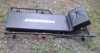 Side view of black Powerbuilt auto repair creeper showing six wheels and padded body with white logo.