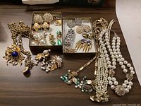 Photo showing assorted costume jewellery including gold-tone lion head necklace, faux pearl bead necklaces, and multiple pairs of rhinestone and bead earrings arranged on wood surface.