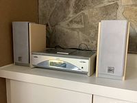 Full view of JVC mini bookshelf stereo system with two speakers on either side, placed on white shelf against textured wall.