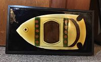 Front view of one ceramic fish tile framed in wood, showing yellow, brown, green, and black colors with some distress on frame corners.