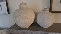 Two round clay vessels with removable lids, one larger (13 inches) and one smaller (11 inches), both with textured light beige surface and decorative lids